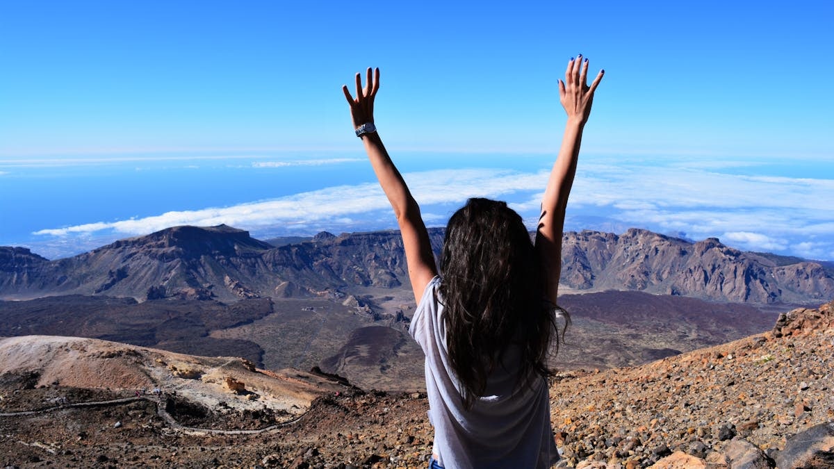 A woman celebrates atop Mount Teide with arms raised, overlooking the vast landscape and sea.